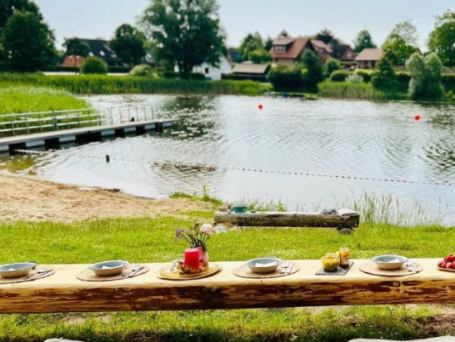 Ein Holztisch mit verschiedenen Speisen an der Badestelle vom Kleinen See Mustin, umgeben von Bäumen.