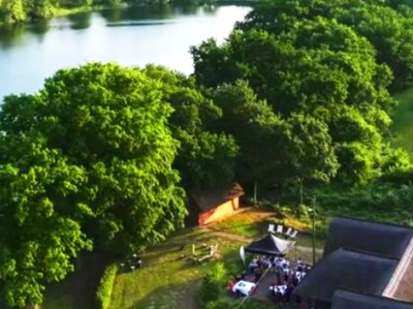 Landschaft mit Fluss und Bäumen, sowie einem Veranstaltungsbereich am Ufer. Haus am See Mustin